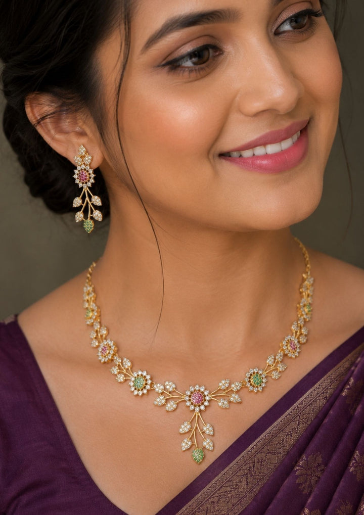 close up offer south indian women wearing Gold-plated zircon diamond necklace and matching drop earrings displayed on a black mannequin bust with dark purple fabric in the background. The jewelry features gold-tone floral and leaf motifs set with clear stones, pink gemstones, and small green accents.