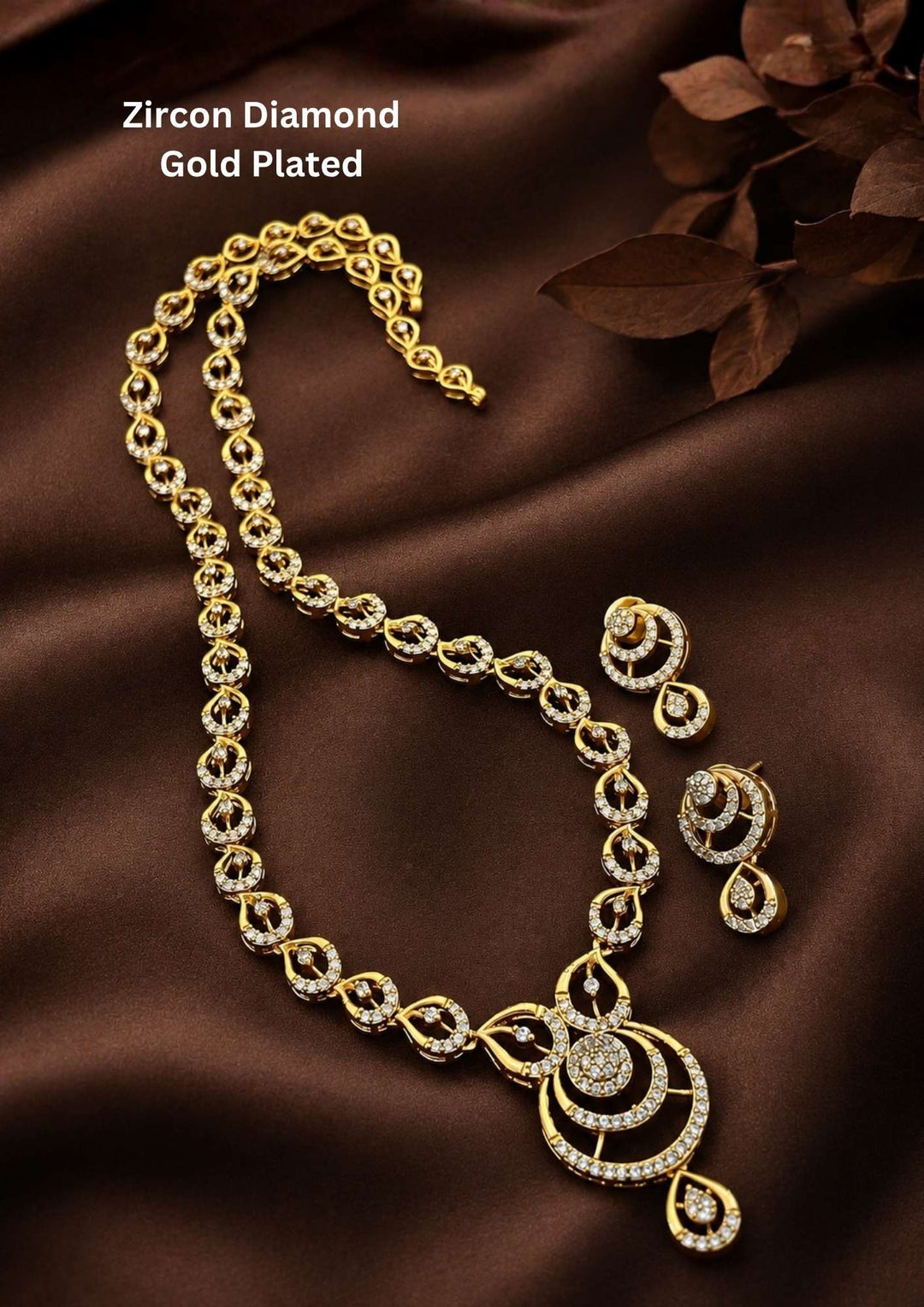 Luxury styling of a cubic zirconia studded gold plated necklace and matching tiered drop earrings displayed next to decorative dried leaves. The high-contrast lighting highlights the fine pavé stone settings and the high-polish yellow gold finish, perfect for festive wedding wear.