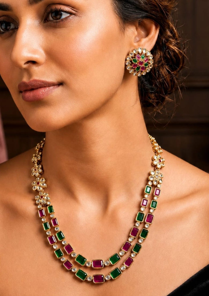 A close-up of a woman wearing a gold-plated double-strand necklace and matching earring set with rectangular green and magenta-red stones.