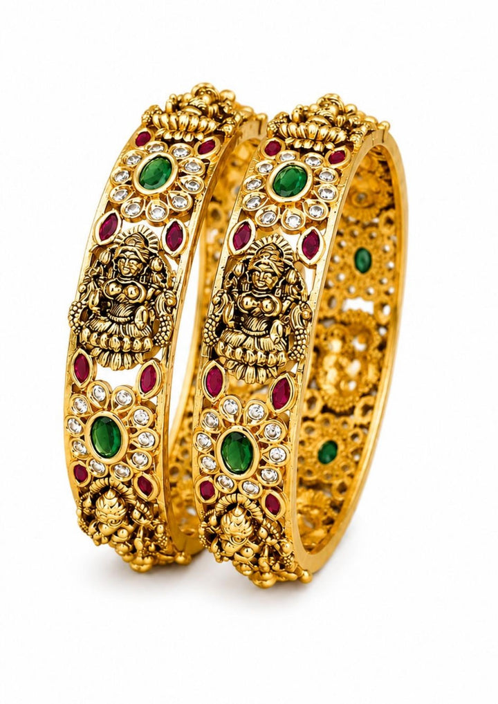 A pair of elegant gold-plated temple bangles featuring beautifully carved motifs of Goddess Laxmi sitting on a lotus. The antique-finish bangles are adorned with oval green emerald-look stones and small ruby-red stone accents, surrounded by tiny sparkling white stones. perfect for traditional wear, festivals, weddings, poojas, functions and gifting