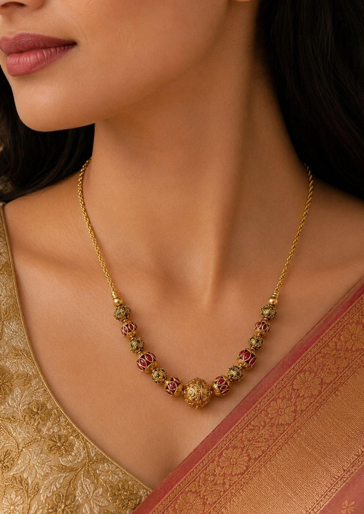 South Indian Women wearing a Gold plated long haraam necklace featuring elegant textured golden spheres alternating with small spacer rings inset with red ruby and green emerald stone . This lightweight chain is crafted with a detailed twisted rope style chain extension held in hand to provide a classy and premium appearance suitable for daily wear, office wear, casual outings, family gatherings, festivals, parties and traditional occasions with skin friendly quality, anti fade polish, comfortable fitting