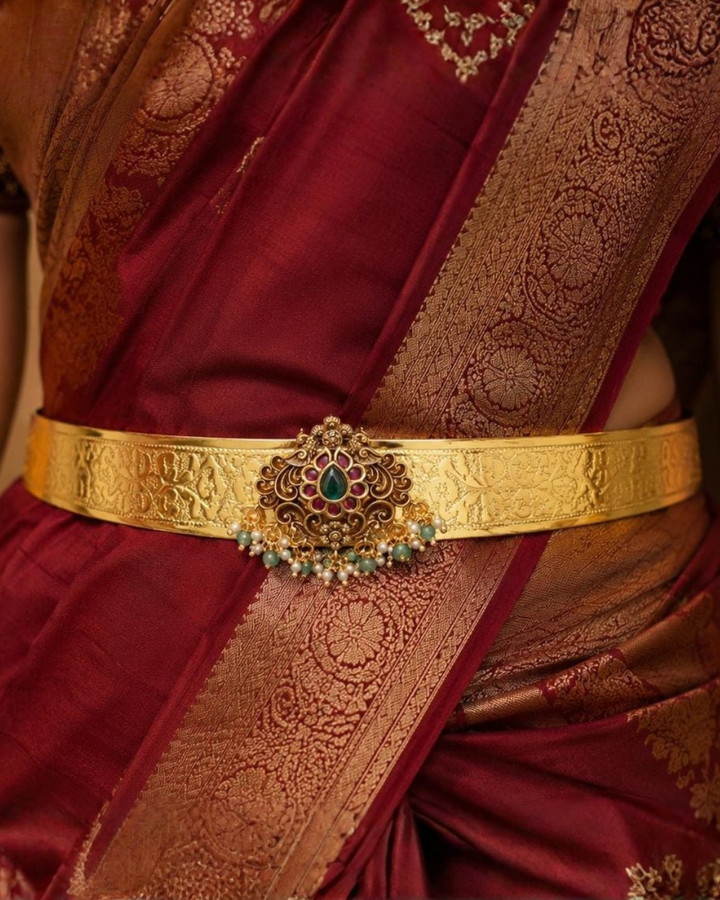 Beautiful gold plated adult hip belt featuring a wide intricately engraved band with a floral gemstone centerpiece decorated with ruby, green stones, and pearl drops. Perfect for traditional wear, weddings, festive occasions, bridal styling, parties, and gifting.