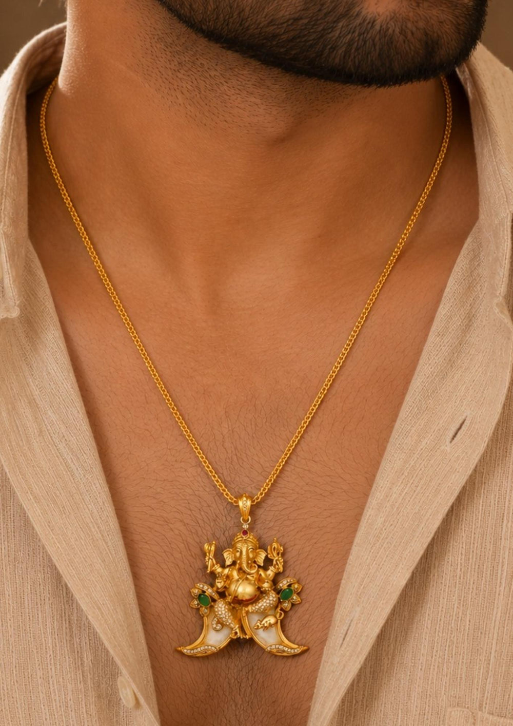 men wearing A traditional gold plated temple style pendant featuring an intricately sculpted idol of Lord Ganesha seated above a curved double tiger claw (pulinakha) motif. The piece is adorned with shimmering white crystals, ruby-red crown details, and green stone side accents, held up by a hand with blurred jewelry store displays in the background.