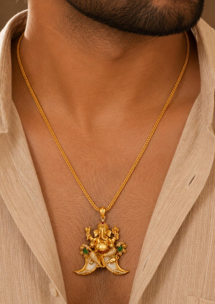 men wearing A traditional gold plated temple style pendant featuring an intricately sculpted idol of Lord Ganesha seated above a curved double tiger claw (pulinakha) motif. The piece is adorned with shimmering white crystals, ruby-red crown details, and green stone side accents, held up by a hand with blurred jewelry store displays in the background.