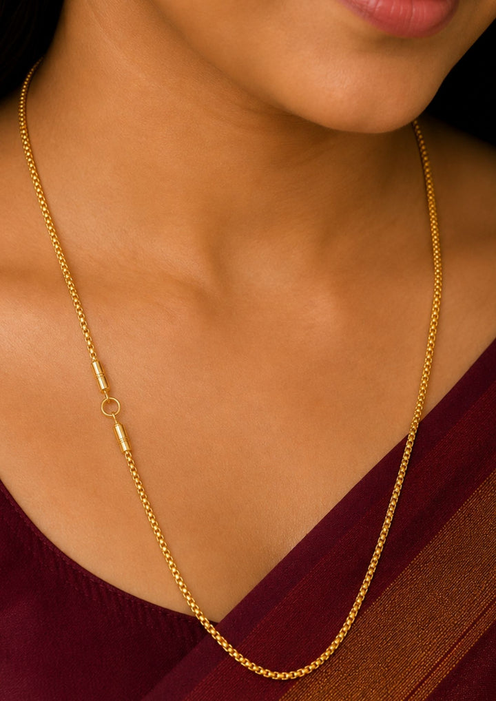 A close-up of a woman wearing a delicate gold chain necklace with a simple cylindrical clasp and ring detail, paired with a deep maroon sari.