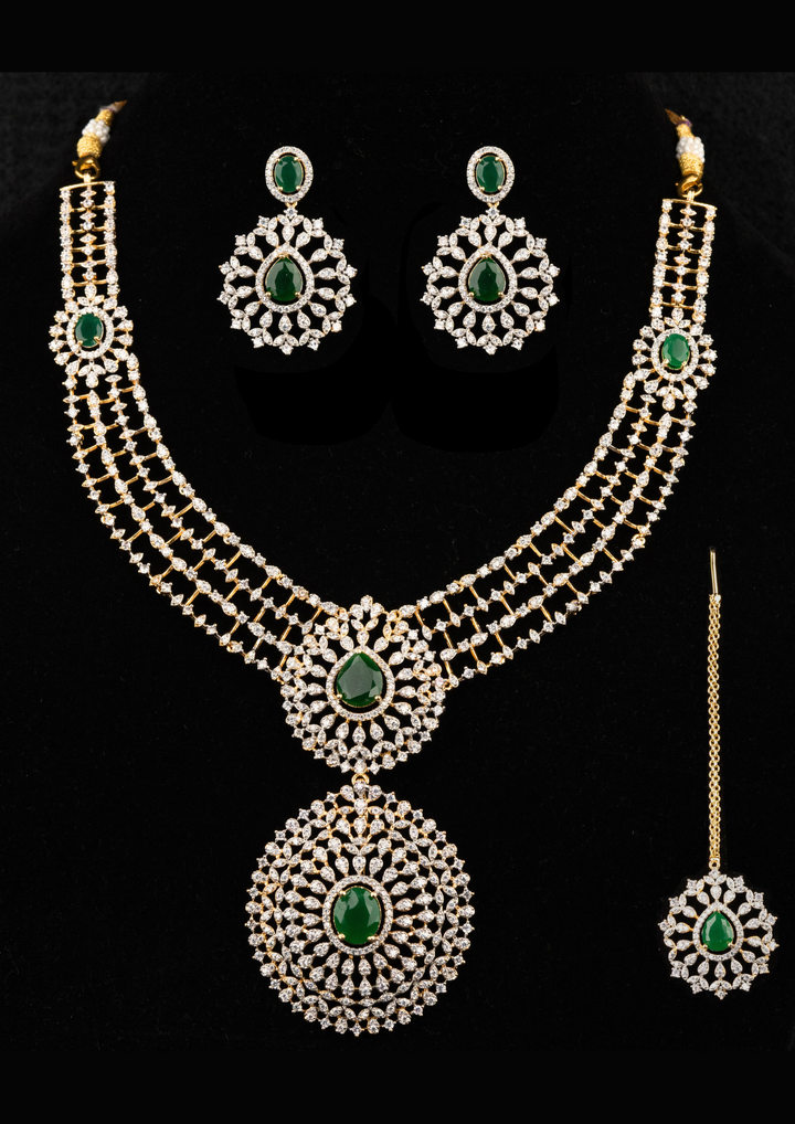 Zircon diamond gold-plated necklace and earrings set designed with 3 row zircon diamonds and in the middle it is designed with green emerald stone and it also has matching earrings designed in the leaf pattern with green emerald stud on the top and it also had magtikka perfect for weddings, bridalwear, festivals and its anti fade color