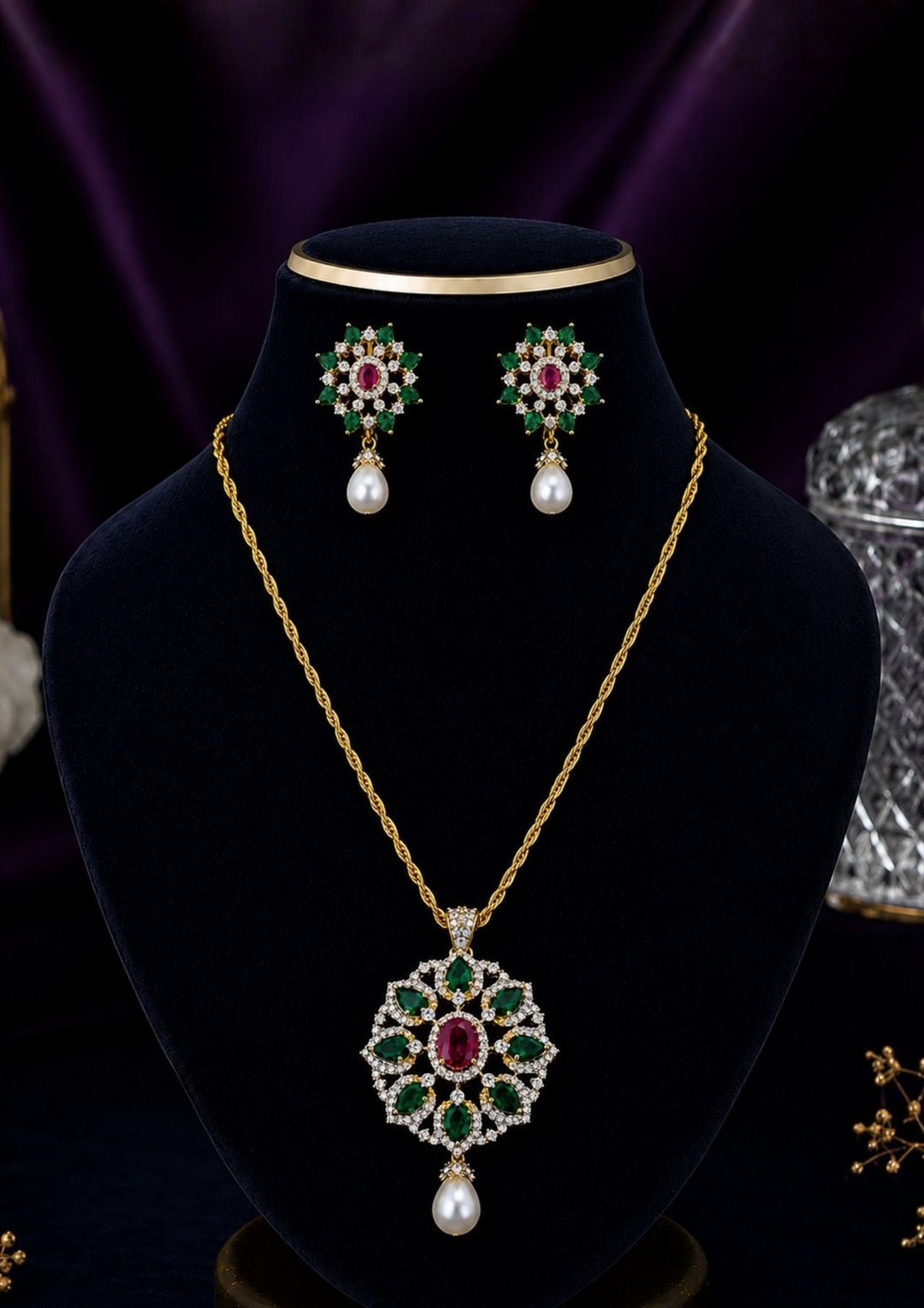 Gold-plated pendant necklace and matching stud earrings displayed on a black velvet jewelry bust. The necklace features a detailed floral pendant with red, green, and blue stones, while the matching earrings are shown above the bust.