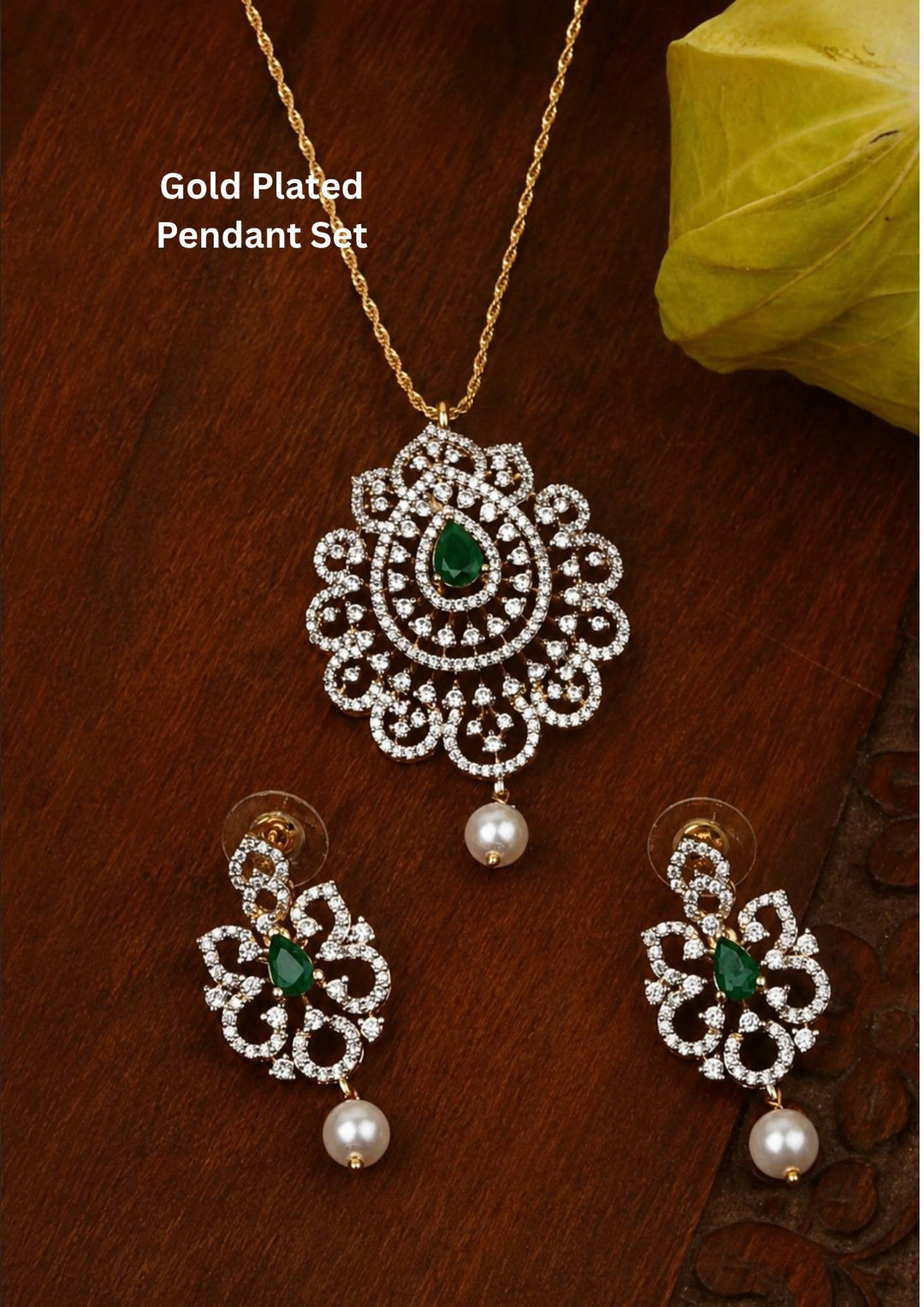 Elegant zircon diamond gold plated pendant set featuring a floral-inspired pendant designed with sparkling white zircon stones arranged in circular patterns. The centerpiece showcases a pear-shaped emerald red stone surrounded by dazzling zircon stones, finished with a delicate pearl drop. The matching earrings mirror the same floral design with green stone and pearl danglers, Perfect for weddings, festivals, office wear, functions and its anti fade color