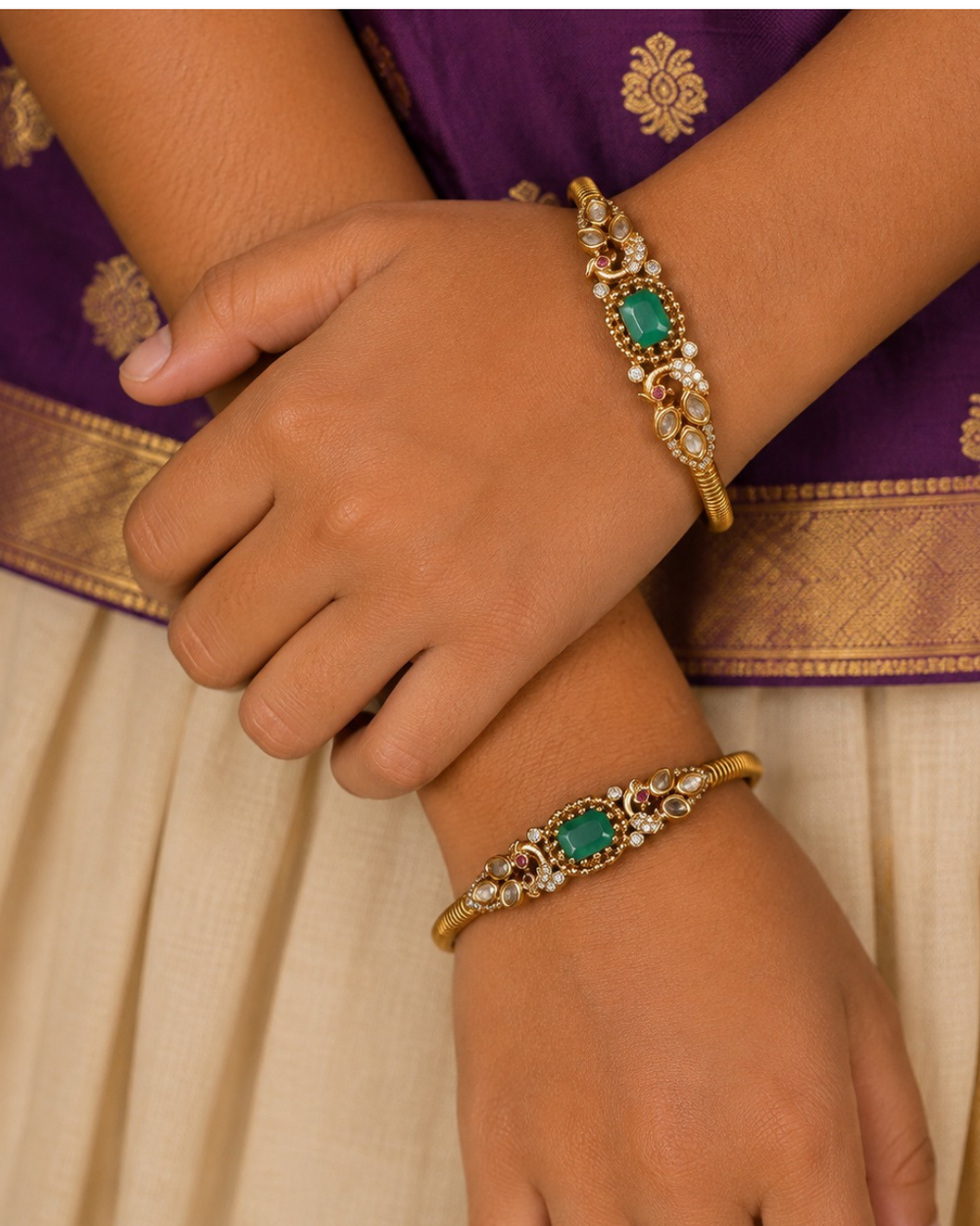 A pair of premium 18k gold plated kada bangles featuring an exquisite twin peacock design flanking a central rectangular emerald green gemstone. The peacock silhouettes are delicately studded with tiny shimmering white stones and accented with ruby red eyes. The remaining portion of the band features a clean, highly polished ribbed texture.