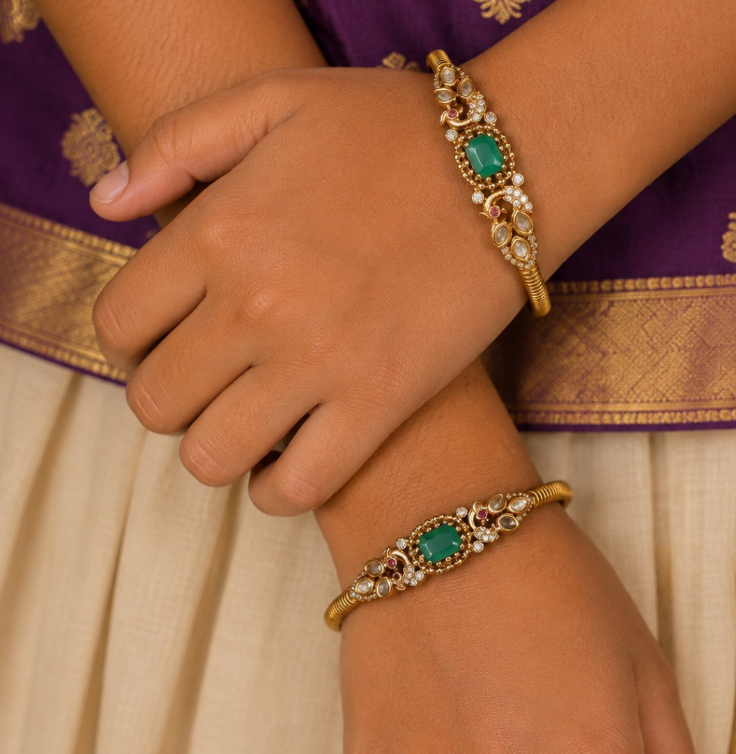 A pair of premium 18k gold plated kada bangles featuring an exquisite twin peacock design flanking a central rectangular emerald green gemstone. The peacock silhouettes are delicately studded with tiny shimmering white stones and accented with ruby red eyes. The remaining portion of the band features a clean, highly polished ribbed texture.