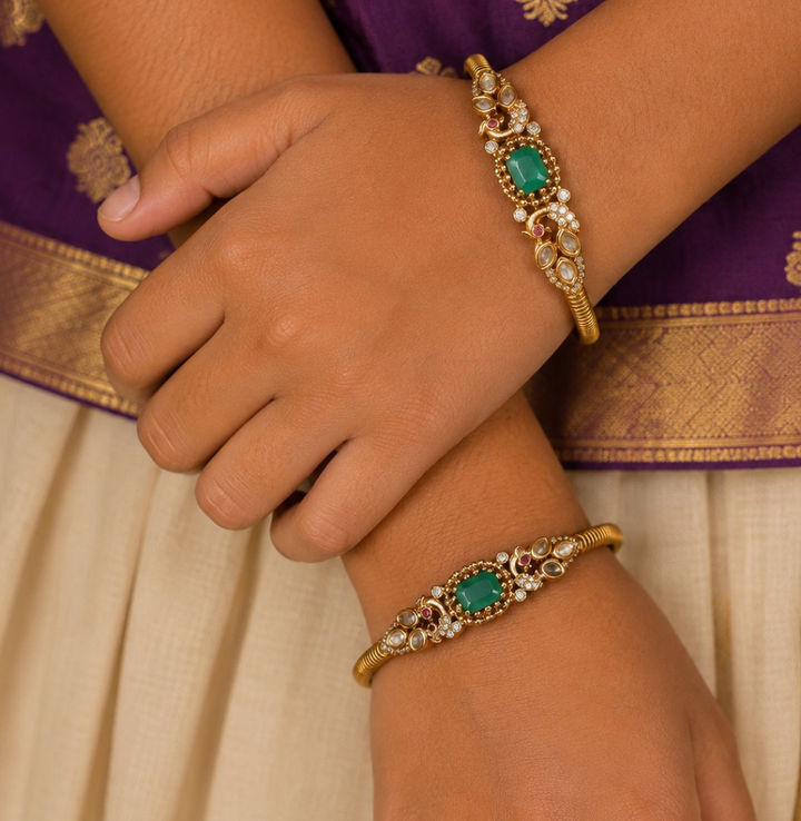 A pair of premium 18k gold plated kada bangles featuring an exquisite twin peacock design flanking a central rectangular emerald green gemstone. The peacock silhouettes are delicately studded with tiny shimmering white stones and accented with ruby red eyes. The remaining portion of the band features a clean, highly polished ribbed texture.