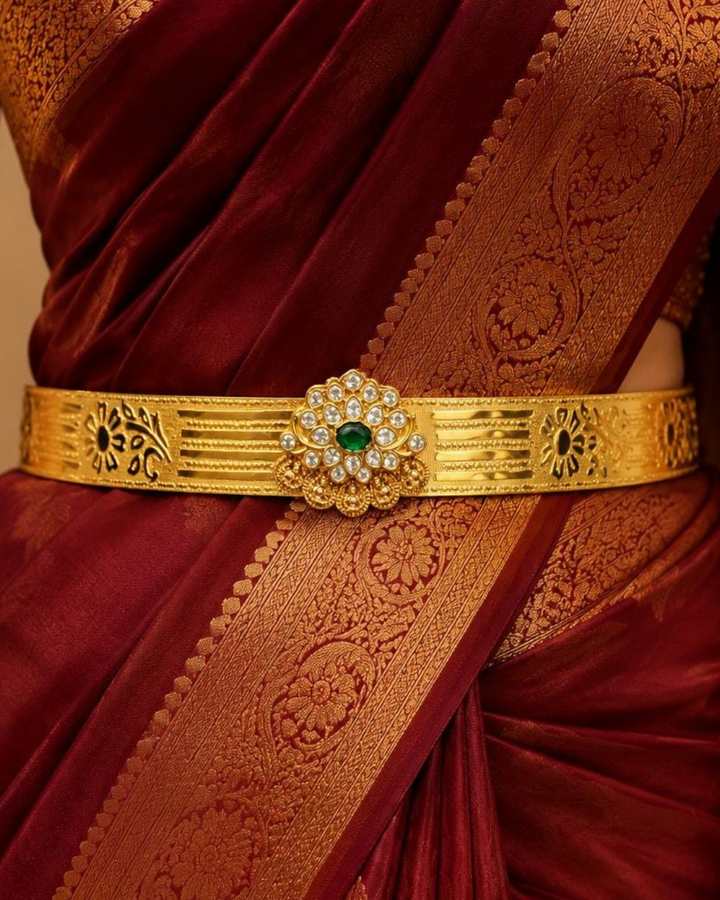 Beautiful gold plated adult hip belt featuring an intricately engraved wide band with floral motifs and a central green gemstone surrounded by sparkling clear stones. Perfect for traditional wear, weddings, festive occasions, bridal styling, parties, and gifting.