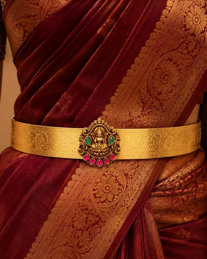 Beautiful gold plated adult hip belt featuring an intricately engraved wide band with a traditional Lakshmi centerpiece decorated with ruby and green stones. Perfect for traditional wear, weddings, festive occasions, bridal styling, parties, and gifting.