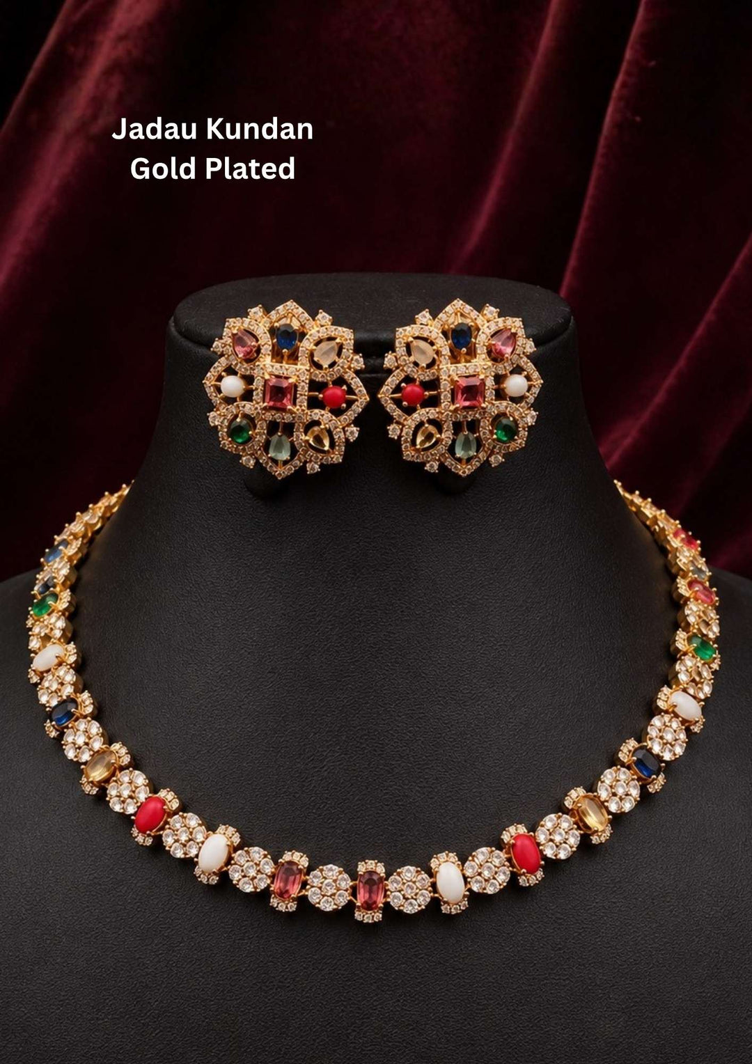 Gold-plated Jadau Kundan necklace and matching stud earrings displayed on a black jewelry bust against a dark red velvet background. The jewelry features gold-toned floral motifs with red, green, blue, and white stones, perfect for parties