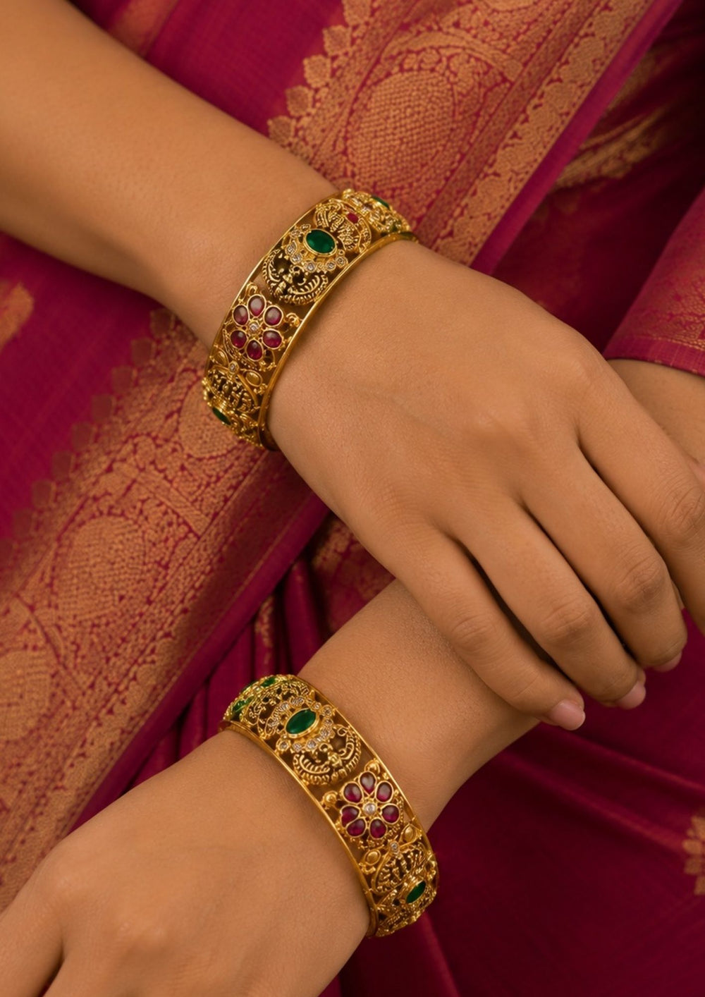 South Indian women wearing A pair of thick Jadau Kundan gold-plated kada bangles featuring intricate traditional motifs. The design showcases detailed peacock carvings, floral patterns inset with ruby-pink and emerald-green stones, and delicate white Kundan accents running along an antique gold-finished frame. perfect for weddings and festive wear.
