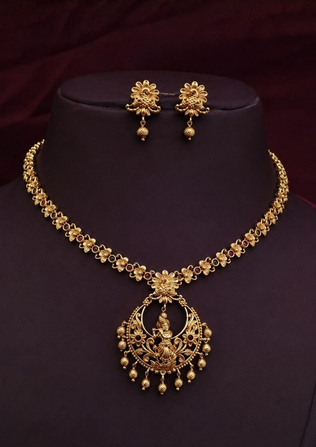 Gold-toned traditional necklace and matching earrings displayed on a dark purple jewelry bust against a dark maroon fabric background.