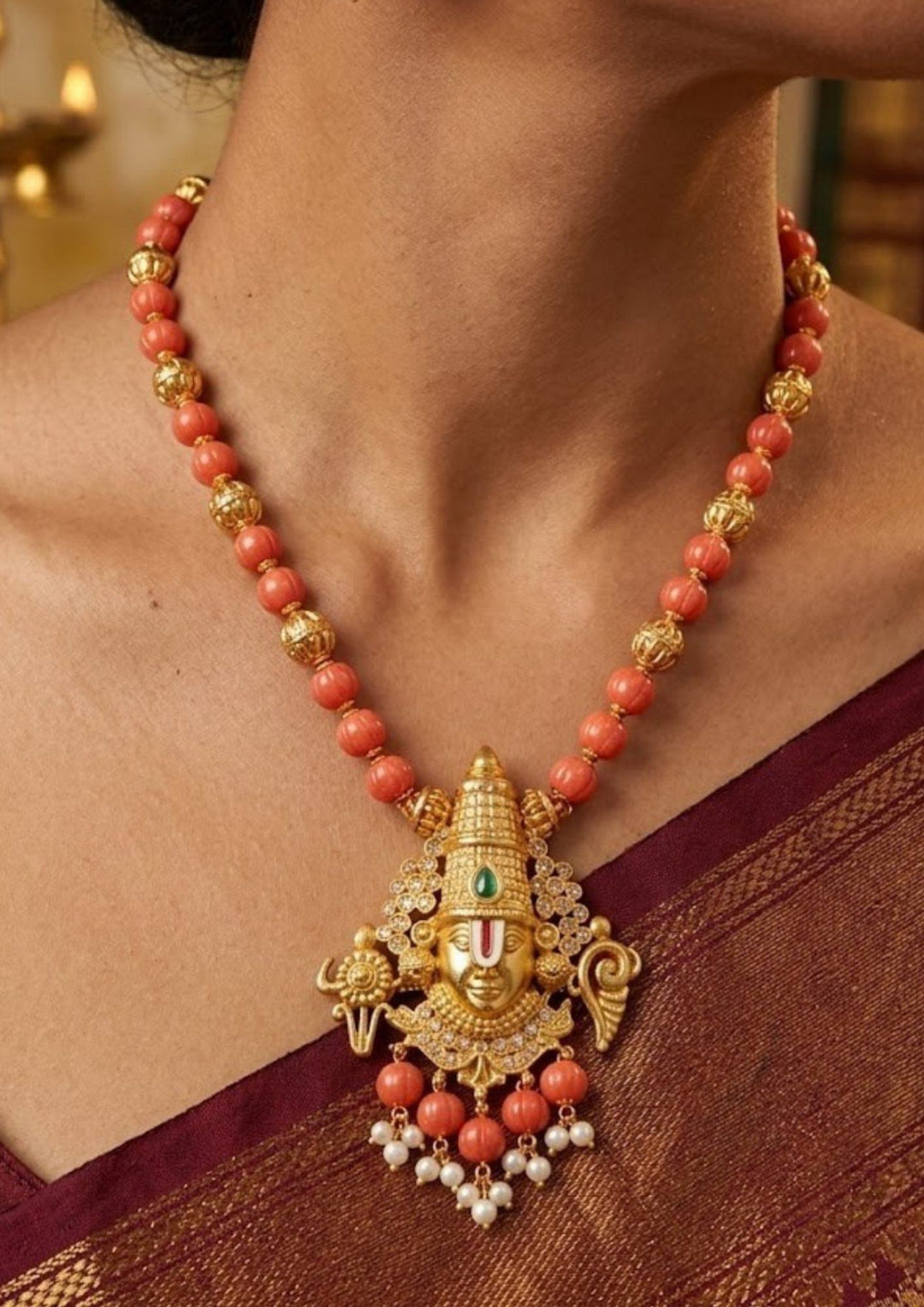 A South Indian woman wearing a Gold Plated Balaji Coral Beads necklace set with matching earring set with a polished gold finish, The design highlights a beautifully carved blalaji pendant suspended from a neat coral bead strand, The long coral mala and matching earrings pattern with Lord Venkateswara designs, pearl drops, guttapusalu and a smooth finish, Perfecr for festivals, poojas, and weddings