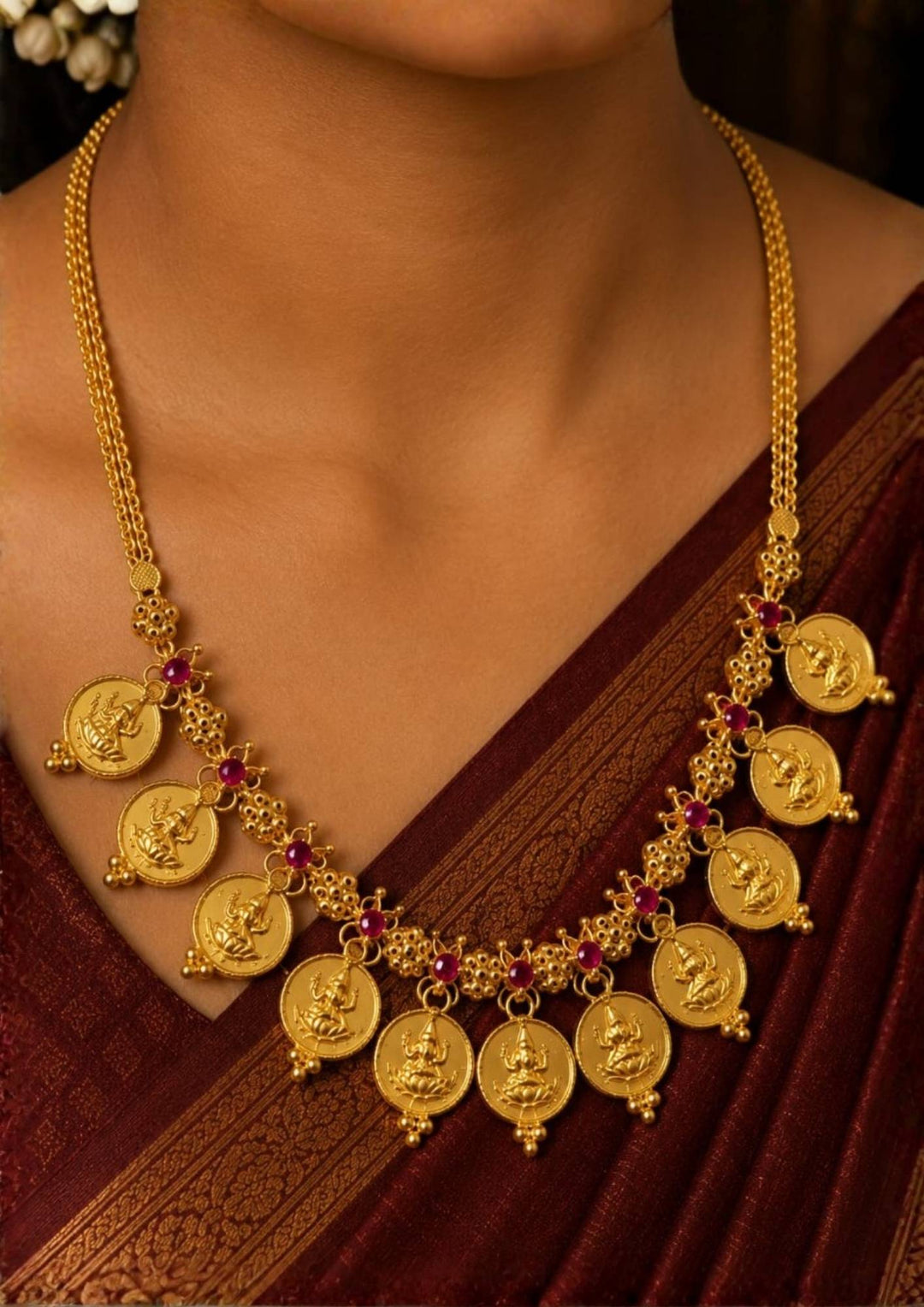 A close-up of a woman wearing a Exquisite Goddess Laxmi gold-plated coin necklace featuring intricate motif carvings and ruby-colored stone accents. Ideal for weddings, festive occasions, temple visits, traditional sarees, and bridal gifting.