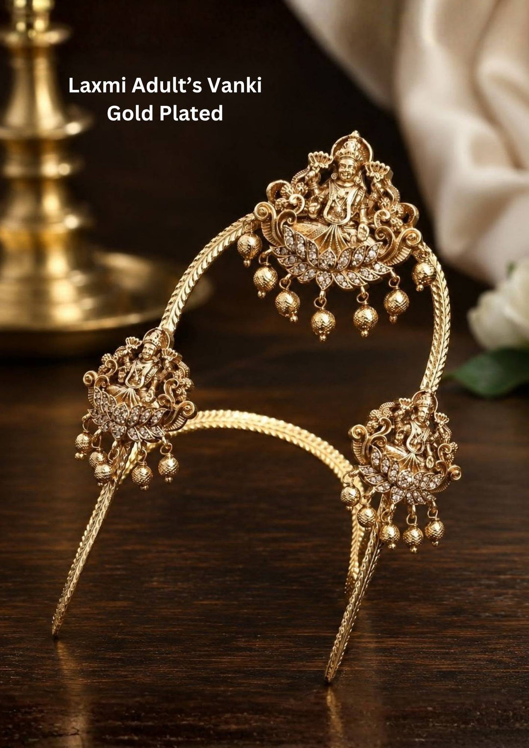 A pair of traditional gold plated temple vanki armlets featuring an elegant V-shaped braided band. Each armlet is adorned with central and side medallions embossed with Goddess Lakshmi seated on a lotus, accented with sparkling white crystals and dangling gold bead drops, displayed against a dark brown fabric background.