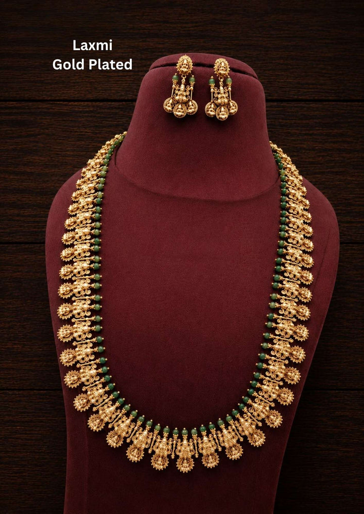 Gold plated long temple haram necklace and matching hanging earrings set featuring a continuous row of traditional textured panels depicting Goddess Laxmi. The inner border of the long chain is decorated with small green beads, and the outer rim is lined with delicate, openwork metal drops. The matching earrings feature small Laxmi coin motifs with hanging bead clusters, providing a premium heritage appearance suitable for festive wear and traditional occasions.