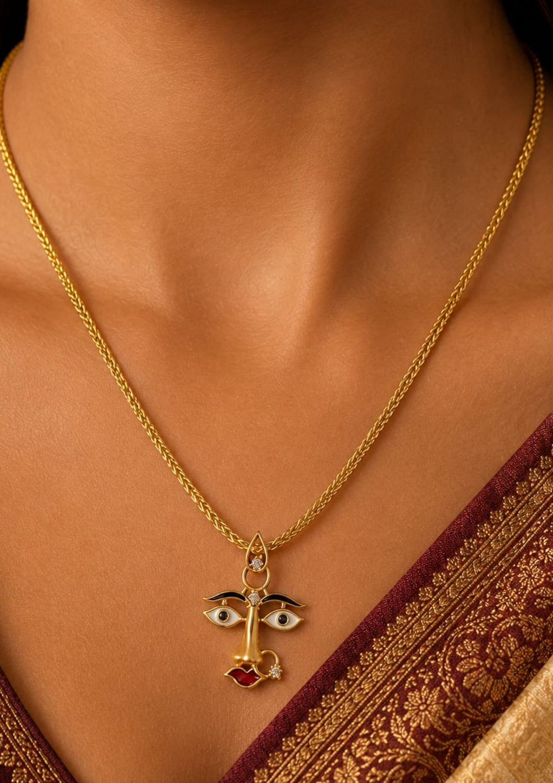 A close-up of a gold chain necklace with a unique pendant featuring two stylized eyes and a small red heart, worn by a model in a traditional maroon and gold saree.