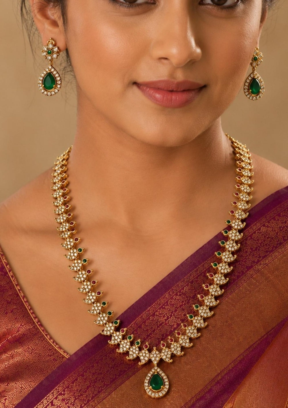  close up offer south indian women wearing Gold-plated traditional Indian jewelry set featuring a necklace and matching earrings with green teardrop-shaped stones and clear stones Premium gold plated long haram featuring sparkling zircon stones with ruby and emerald accents, paired with matching earrings.