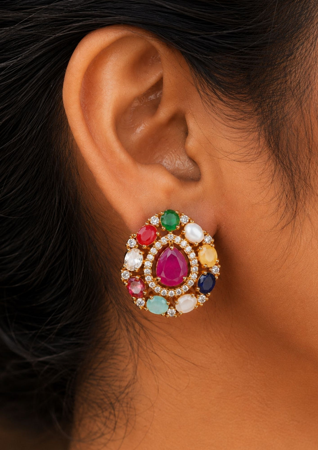 Close-up of an ear wearing a  pair of classic gold plated Oval stud earrings showcasing the traditional Navaratna design with nine distinct multi-colored gemstones. The center holds a smooth oval pink ruby stone, encircled by eight tear-drop shaped petals representing pearl, coral, emerald, sapphire, and other colorful crystals. The set is crafted with a highly polished golden finish and comfortable post backings.