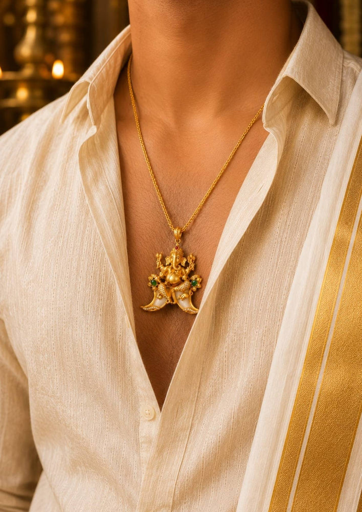 men wearing A traditional gold plated temple style pendant featuring an intricately sculpted idol of Lord Ganesha seated above a curved double tiger claw (pulinakha) motif. The piece is adorned with shimmering white crystals, ruby-red crown details, and green stone side accents, held up by a hand with blurred jewelry store displays in the background.