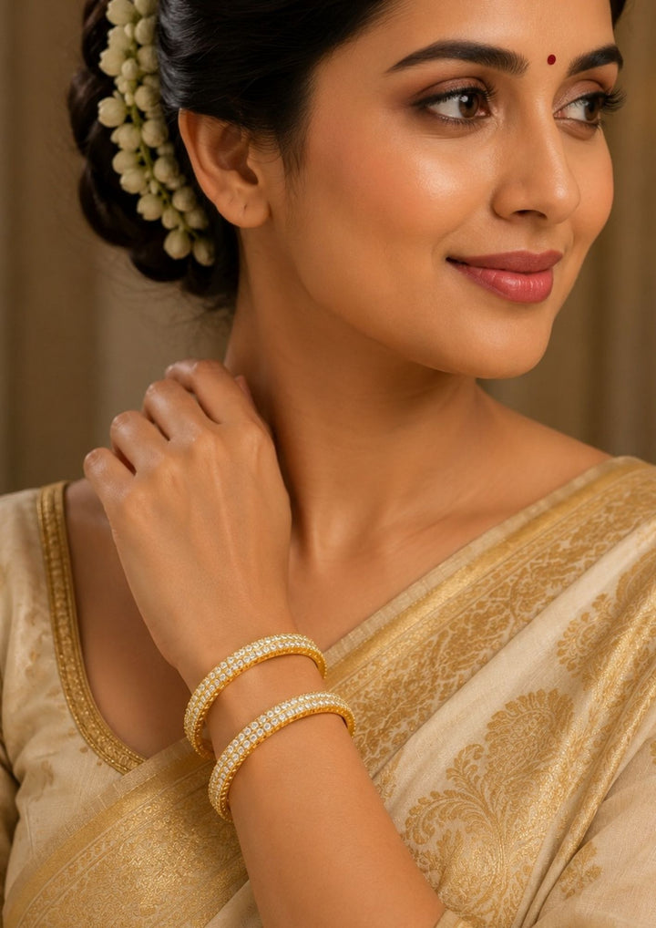 South indian women wearing A pair of premium gold-plated bangles meticulously studded with two parallel rows of brilliant, round-cut clear zircon stones. Each crystal is secured in a classic prong setting along the outer rim of the band, creating a continuous, shimmering diamond-like effect against the rich, polished gold finish suitable for weddings, poojas and bridal occasions.