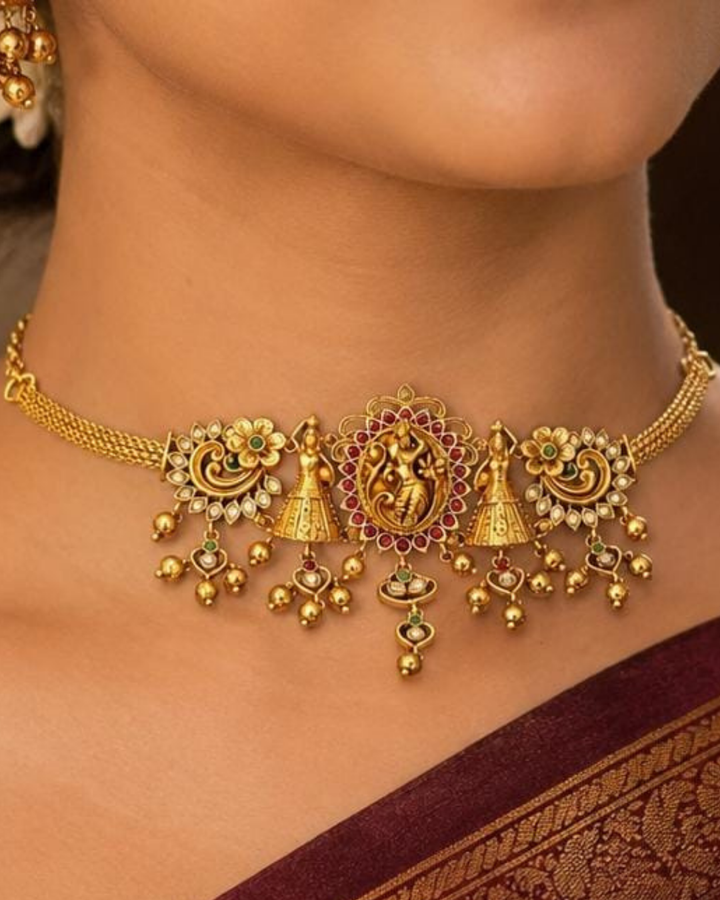 Beautiful gold plated 2-in-1 choker and bajuband set featuring Goddess Lakshmi, peacock and floral motifs embellished with green and pink stones and delicate hanging gold beads. Perfect for weddings, festive occasions, traditional wear, Bharatanatyam styling, parties and gifting.