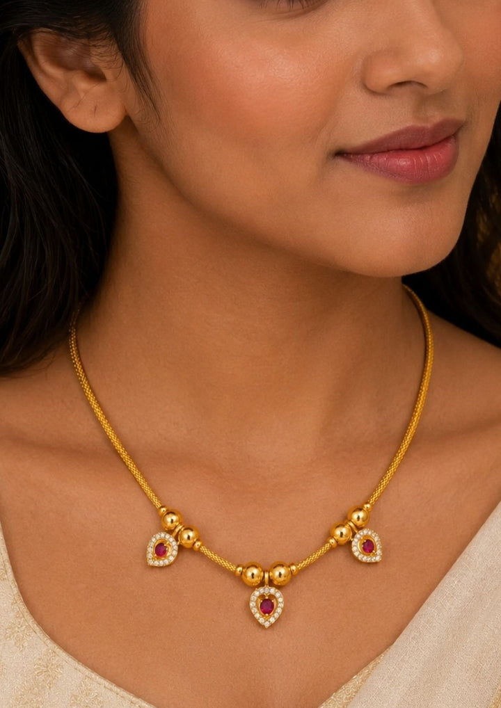 Close-up of a woman wearing a A sleek and minimalist 18k gold plated festive chain necklace featuring a delicate woven rope link. The chain is interspersed with small, polished gold spheres that lead to three hanging heart-shaped drop charms encrusted with clear white zircon diamonds and central vibrant ruby-red stones, perfect for weddings, dailywear, functions comes with anti fade color and skin friendly