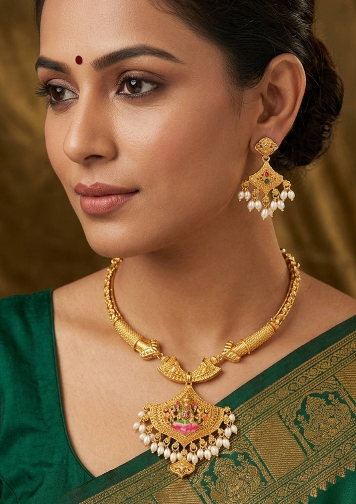 South Indian Woman wearing A traditional temple-style gold-plated choker necklace set featuring Goddess Laxmi. The central fan-shaped pendant beautifully displays a seated Goddess Laxmi motif accented with pink and green enamel work, connected to a textured rigid collar chain and finished with elegant hanging white pearl drops. Perfect for for bridal and festive wear. It is anti tarnish and skin friendly