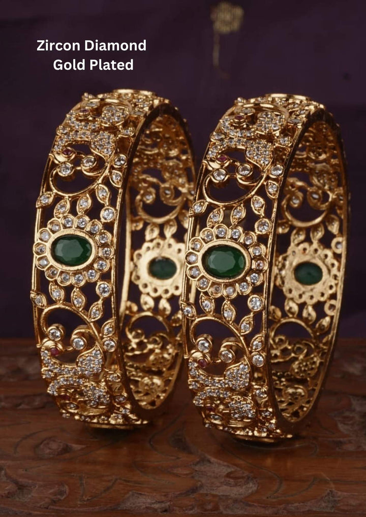 Gold Plated Emerald Bangles with oval green emerald-style stones placed between sparkling zircon diamond detailing and traditional gold pattern work. The bangles feature rich bridal-style design, shiny gold plated finish, lightweight comfort, sweat proof quality, anti-fade colour, smooth edges, skin-friendly finish, and are suitable for weddings, receptions, pooja functions, festivals, and traditional jewelry collections.